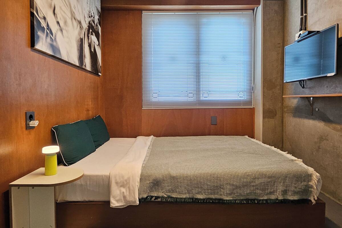 Ett sovrum med en säng, ett sängbord med en lampa, ett fönster med persienner och en tv som är monterad på väggen.