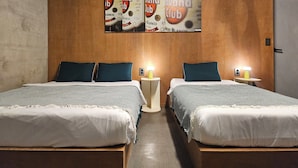 Habitación de hotel con dos camas, cada una con una colcha a rayas blancas y grises, almohadas color turquesa y un cabecero de madera. Hay dos mesitas de noche con lámparas, y un cartel grande de ron 'Cuban Club' en la pared.