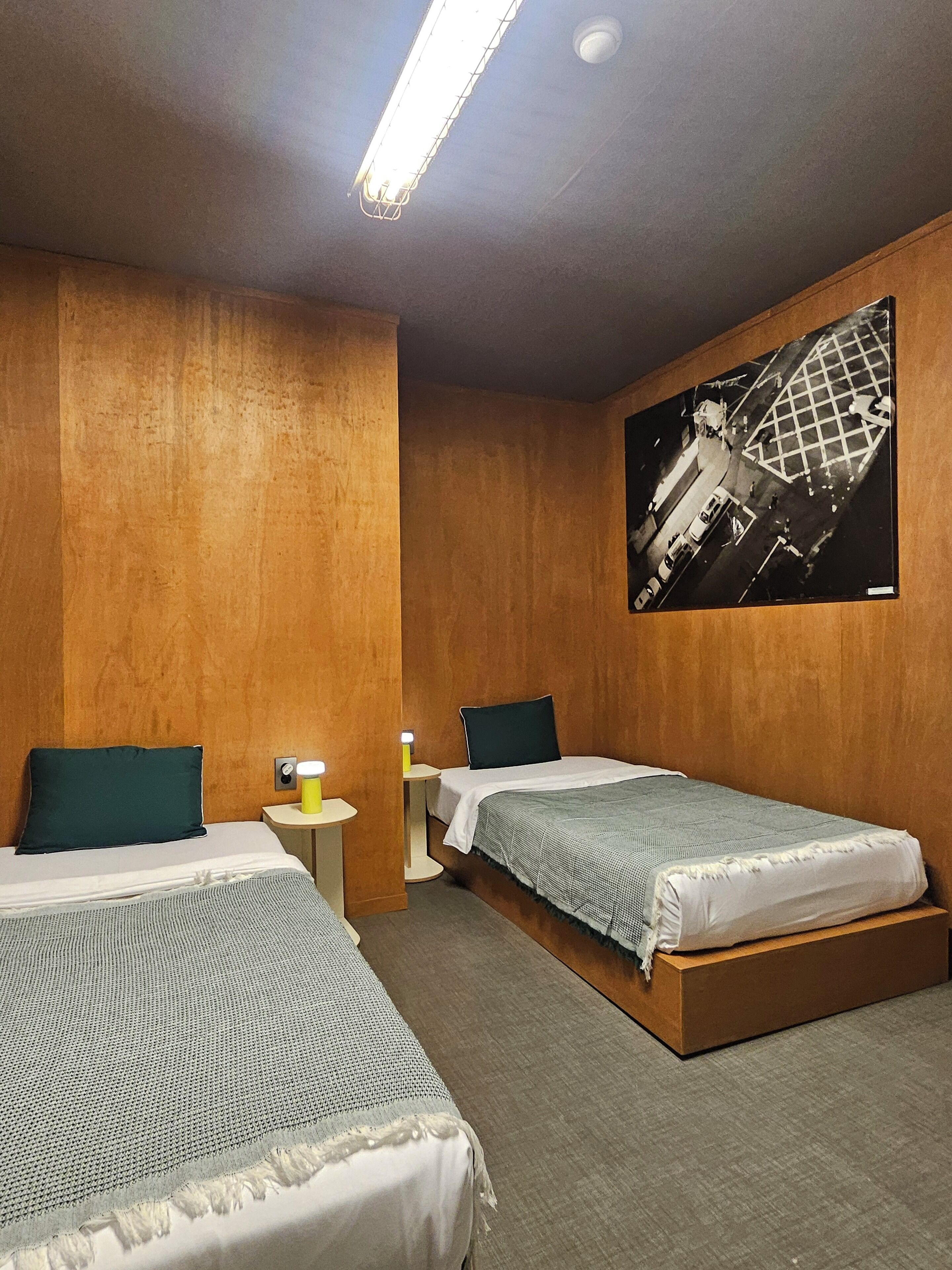 A hotel room with two single beds, wooden walls, a large black and white photo on the wall, and a ceiling light.