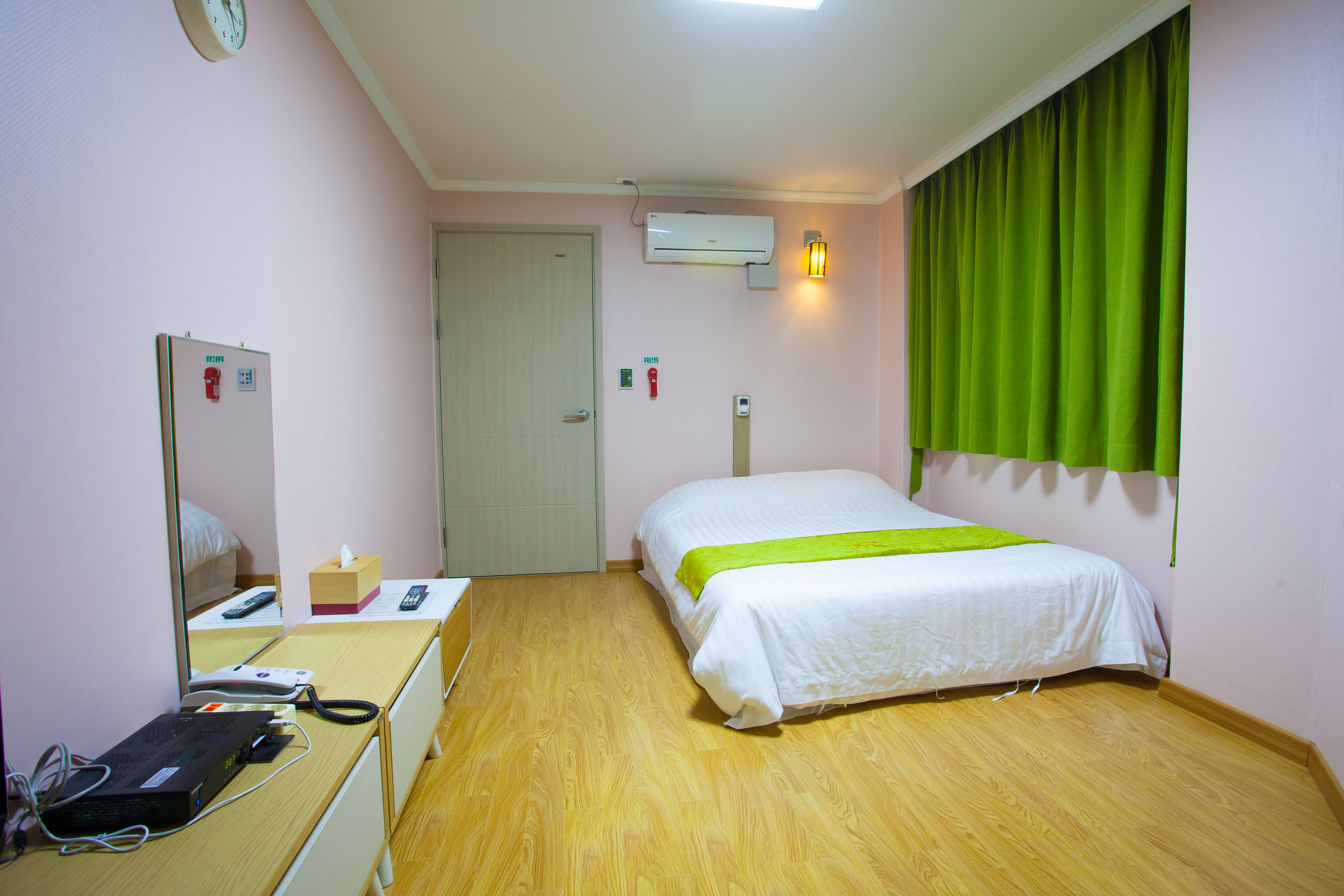 Une chambre d’hôtel avec un lit, un bureau équipé d’un ordinateur, un miroir, un climatiseur et un rideau vert.