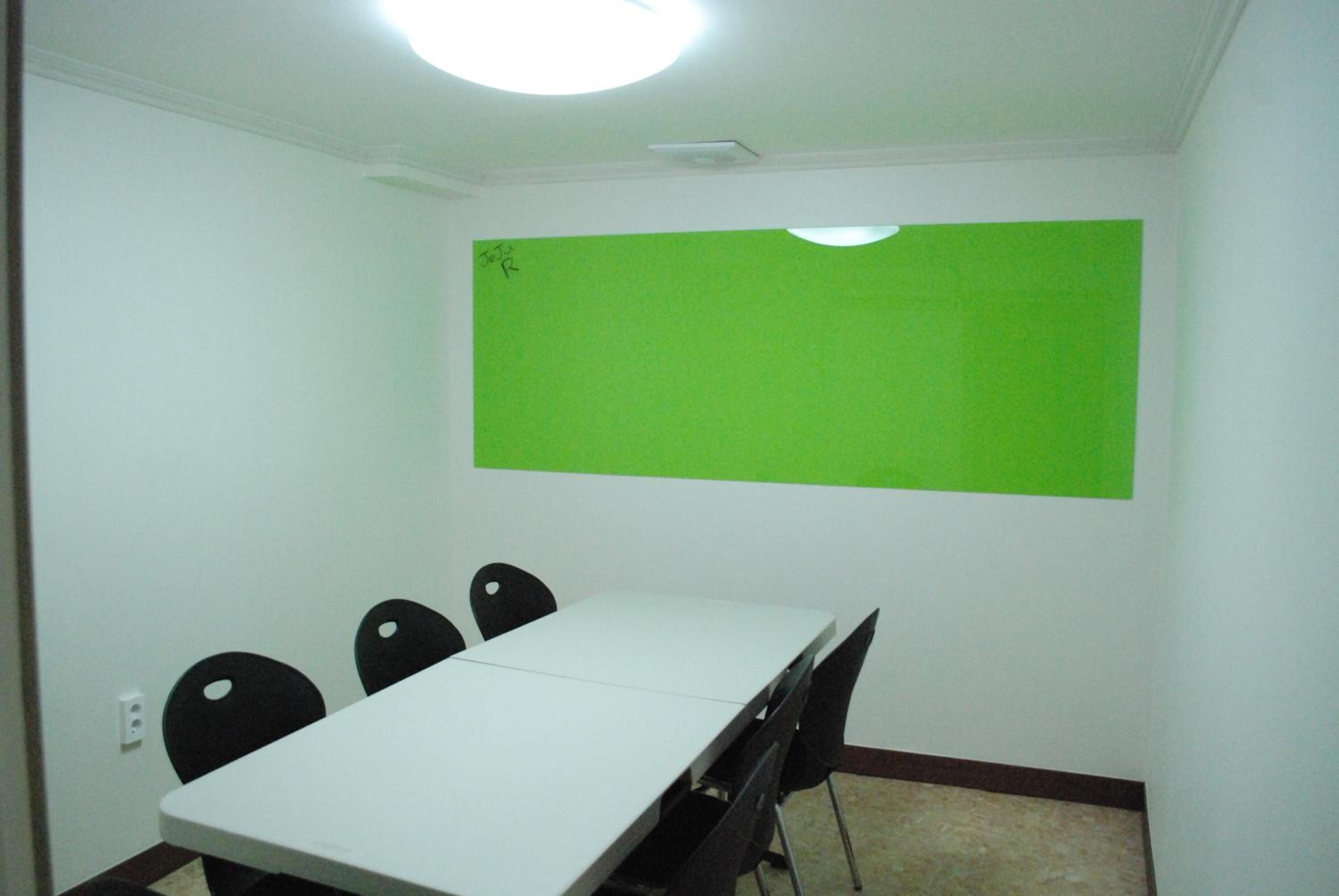 A conference room with a white table, four black chairs, and a green screen on the wall.