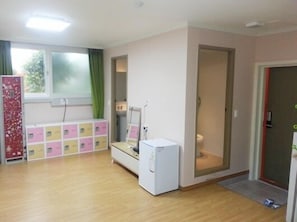 Une petite pièce lumineuse, avec un plancher en bois, un réfrigérateur blanc, un lit doté d’une tête de lit blanche et une fenêtre ornée de rideaux verts.