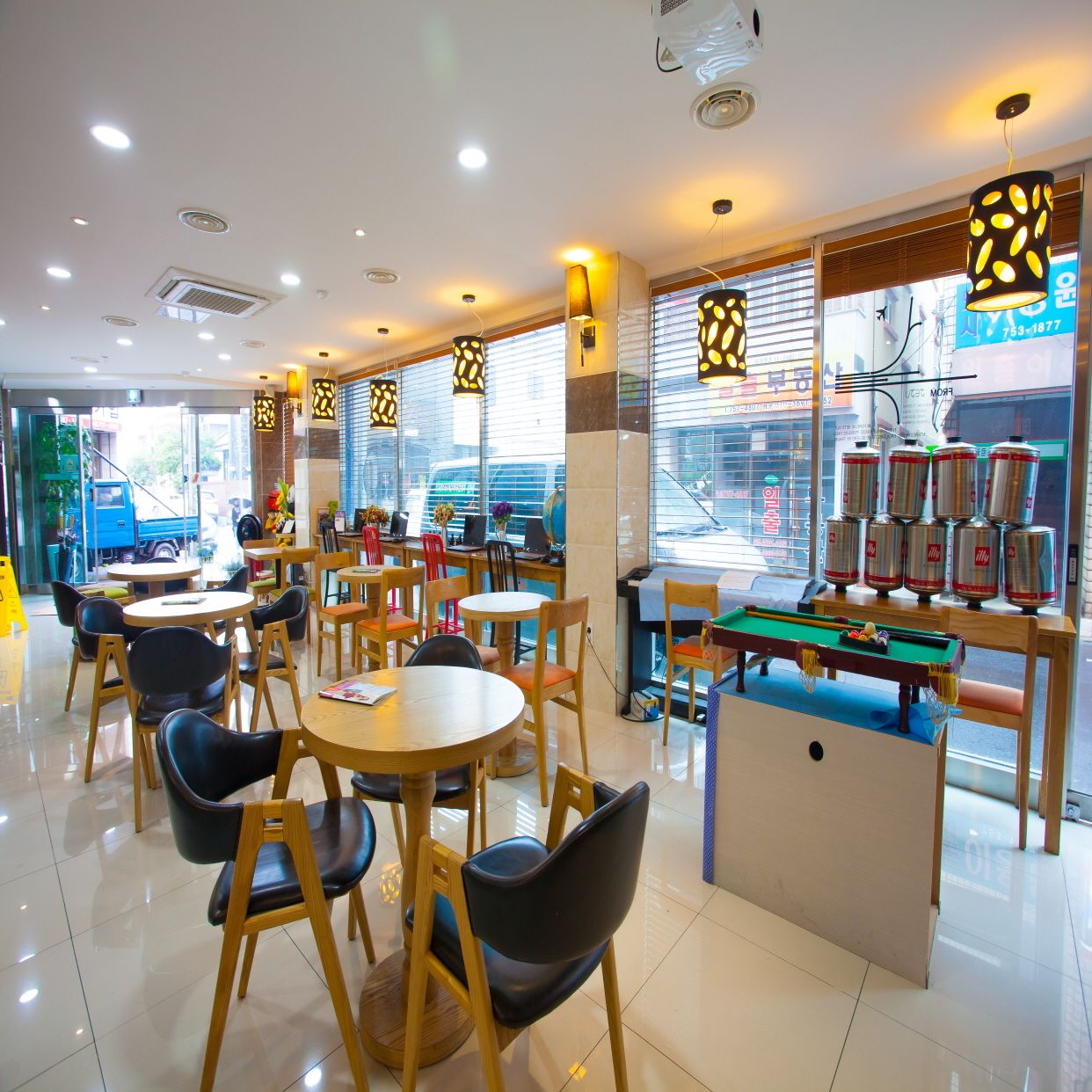 A well-lit indoor space with round tables and chairs, a pool table, and a display of canned beverages.