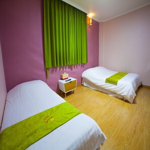 Een hotelkamer met twee bedden, houten vloeren, een nachtkastje met een zakdoekendoos en een groen gordijn.