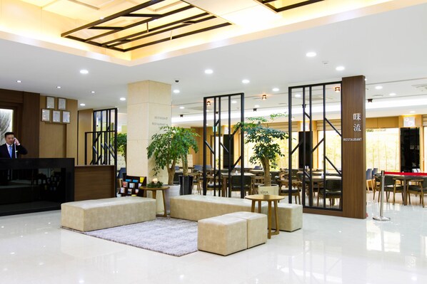 Un hall d’hôtel moderne avec un comptoir d’accueil, un espace salon et une enseigne indiquant la présence d’un restaurant.