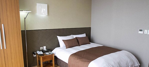 A hotel room with a bed, bedside table, lamp, and wall-mounted thermostat. - Black Sands Hotel (Jeju City)