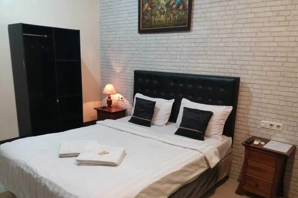 Una cama bien hecha con sábanas blancas y almohadas negras, una mesita de noche con lámpara y un pequeño tocador de madera con un papel encima.