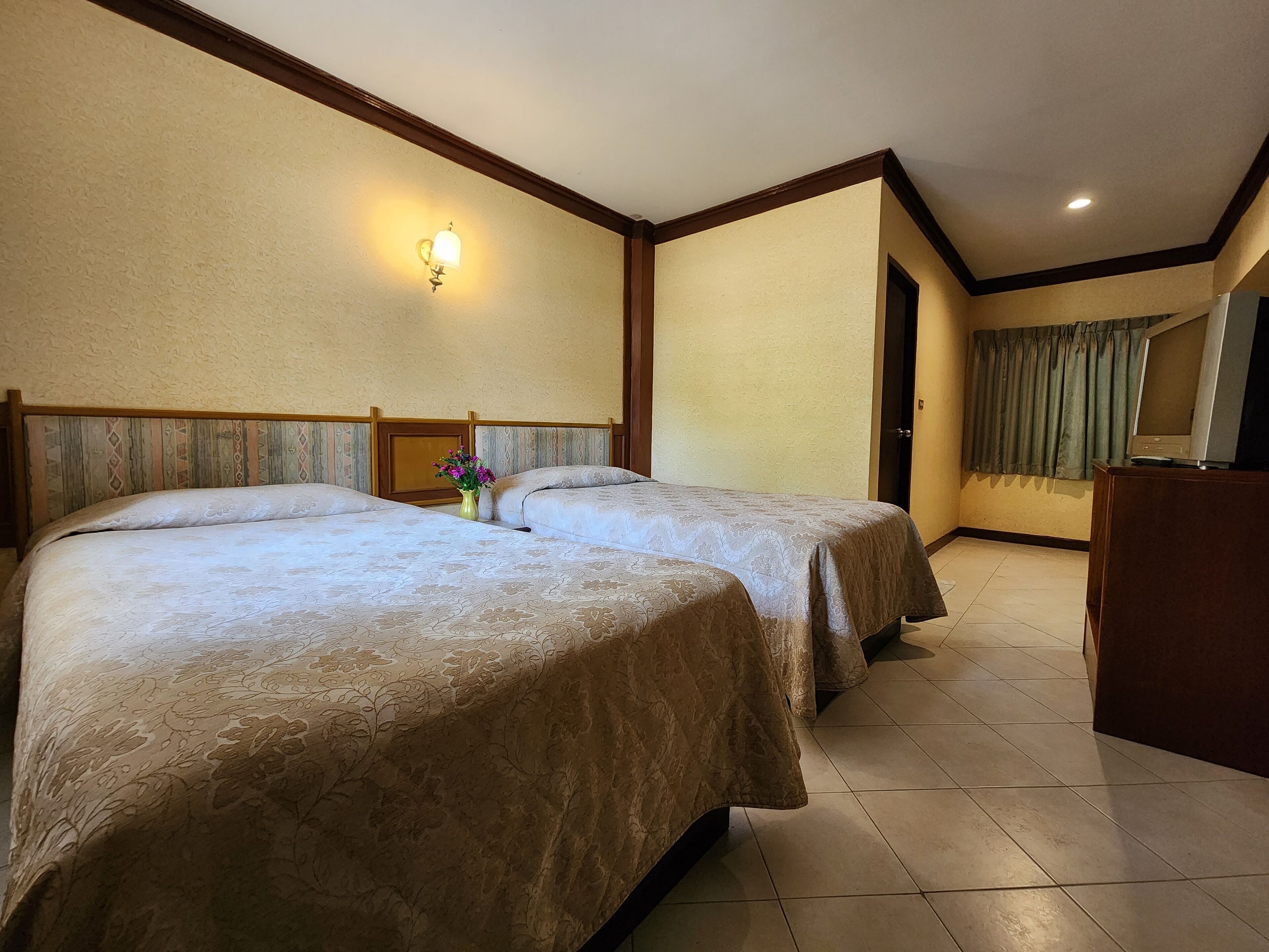 A hotel room with two beds, a nightstand, a vase with flowers, a television, and a window with curtains.