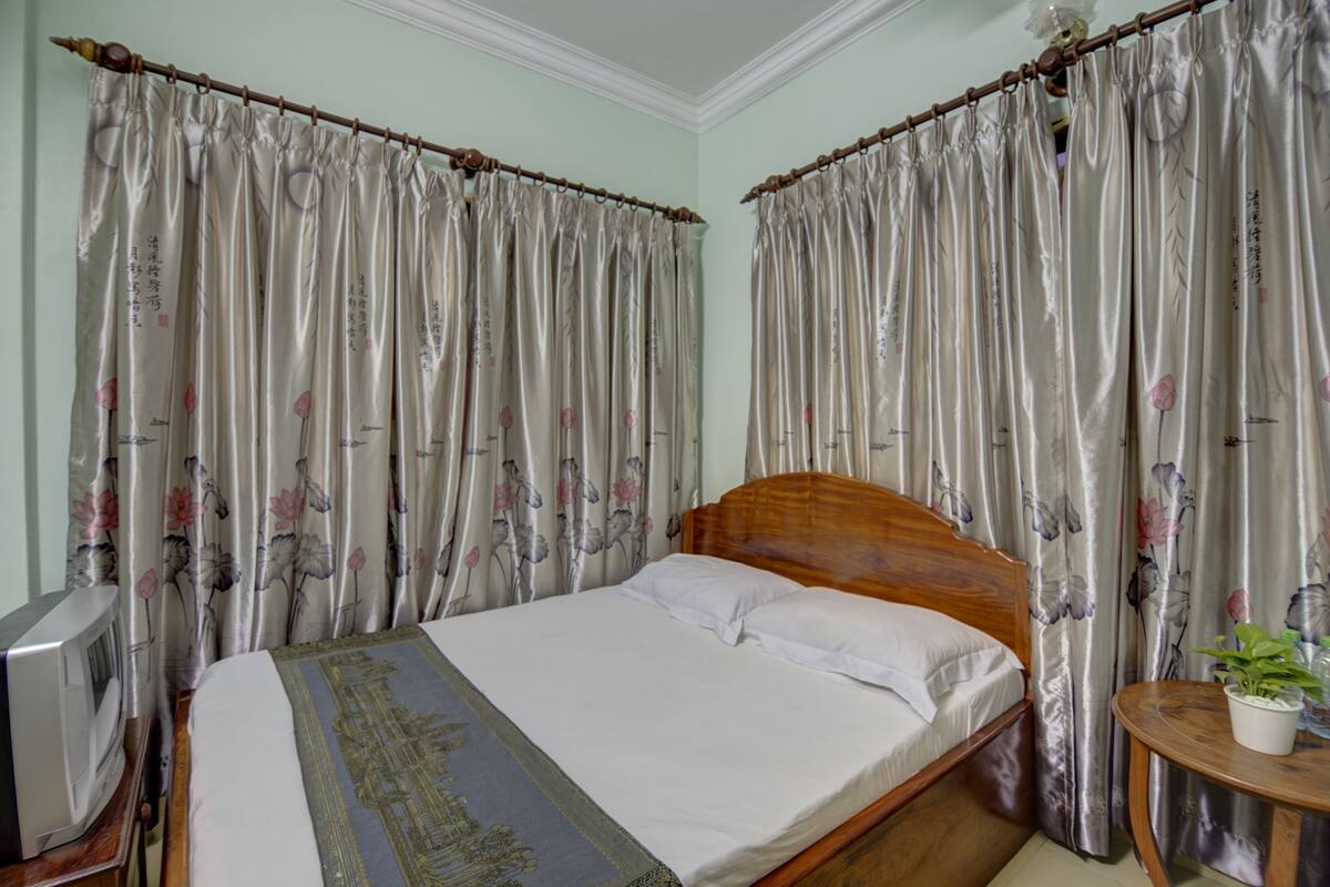 Une chambre à coucher avec un lit en bois, un téléviseur posé sur un meuble en bois et des rideaux  à motifs floraux.