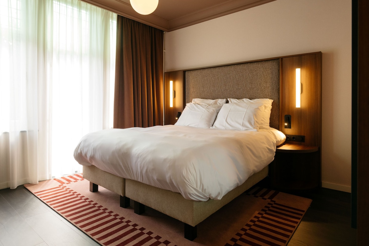 A hotel room with a large bed, a headboard with integrated lighting, a bedside table, and a window with curtains.