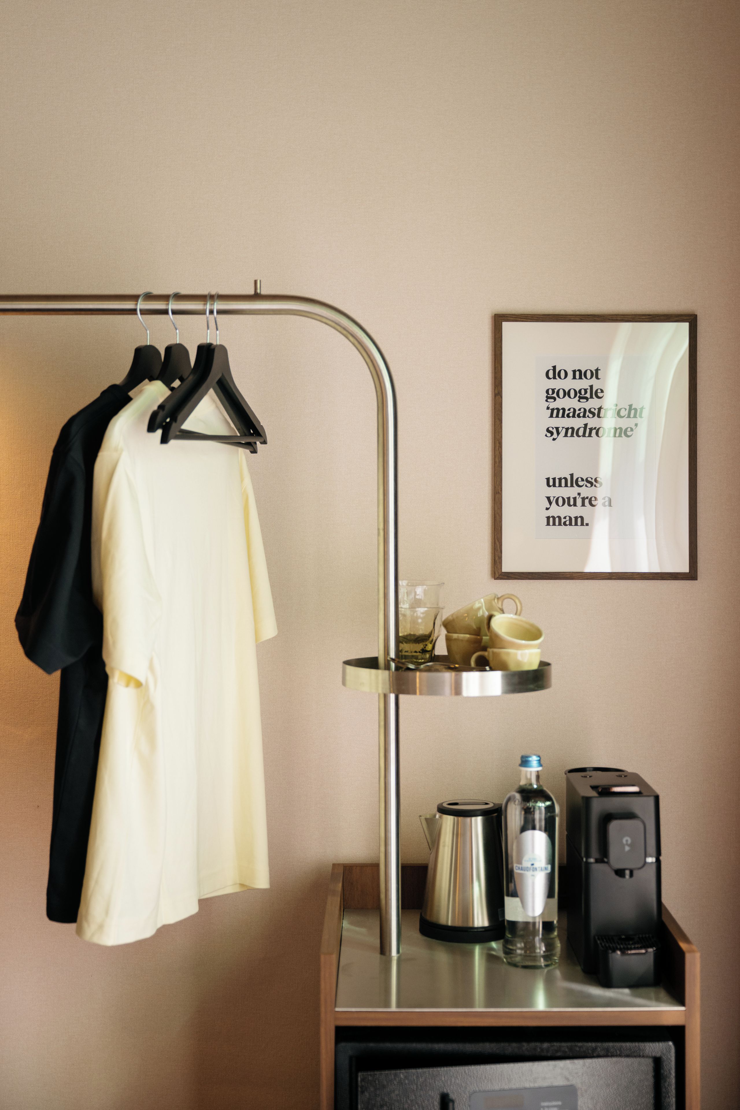 Habitación de hotel con un perchero que sostiene una chaqueta negra y una camisa blanca, una cita enmarcada en la pared, una bandeja con tazas y una cafetera, y un altavoz negro sobre un mueble de madera.