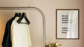 A hotel room with a coat rack holding a black jacket and a white shirt, a framed quote on the wall, a tray with cups and a coffee pot, and a black speaker on a wooden cabinet.