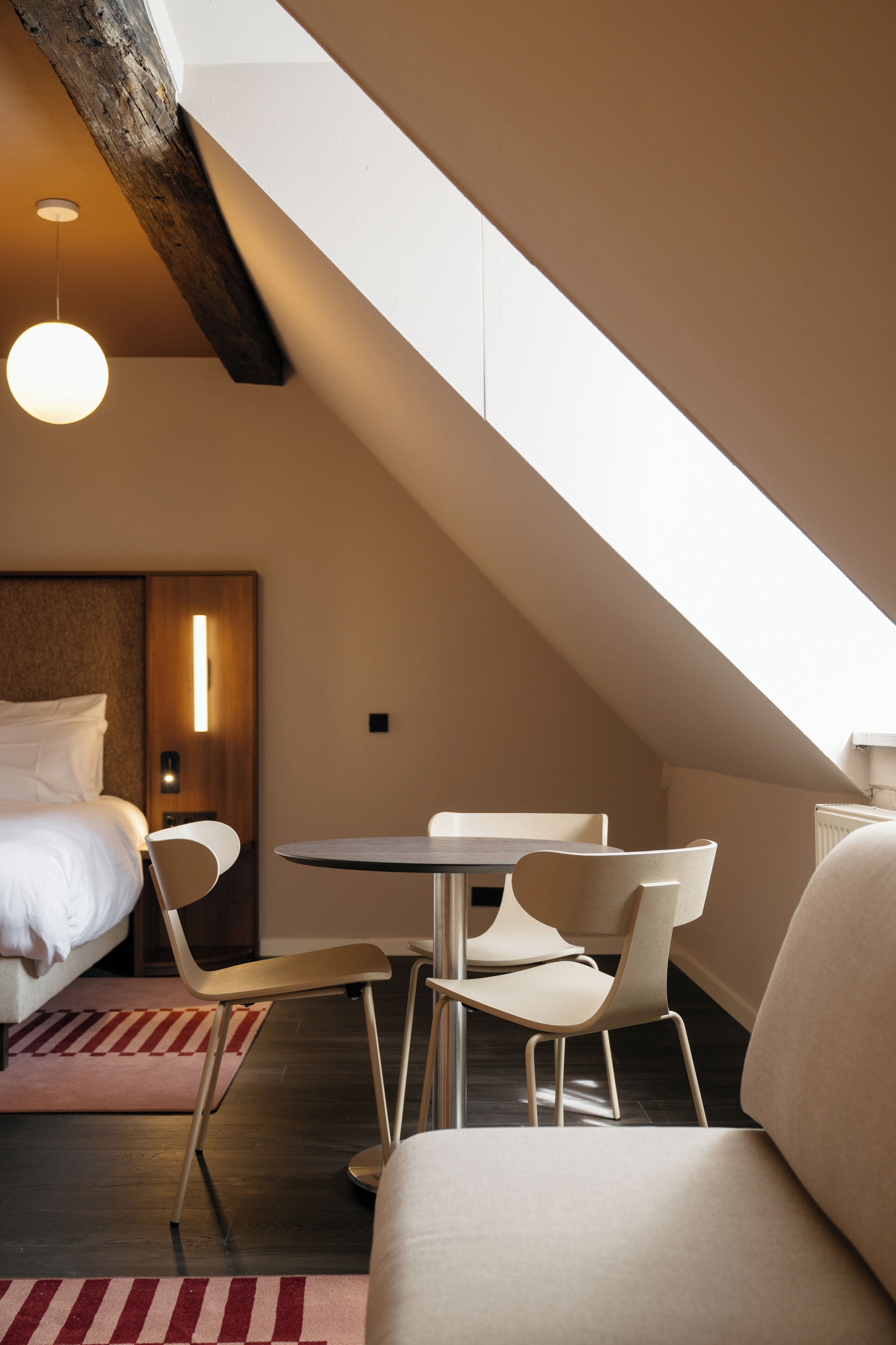 A modern hotel room with a sloped ceiling, a bed, a round table, two chairs, and a sofa.