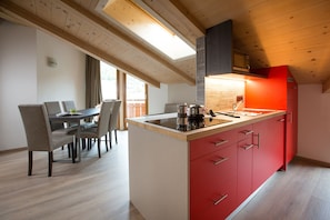 A modern kitchen with a central island, wooden ceiling beams, and a red accent wall. - Appartamenti Isola (Livigno)