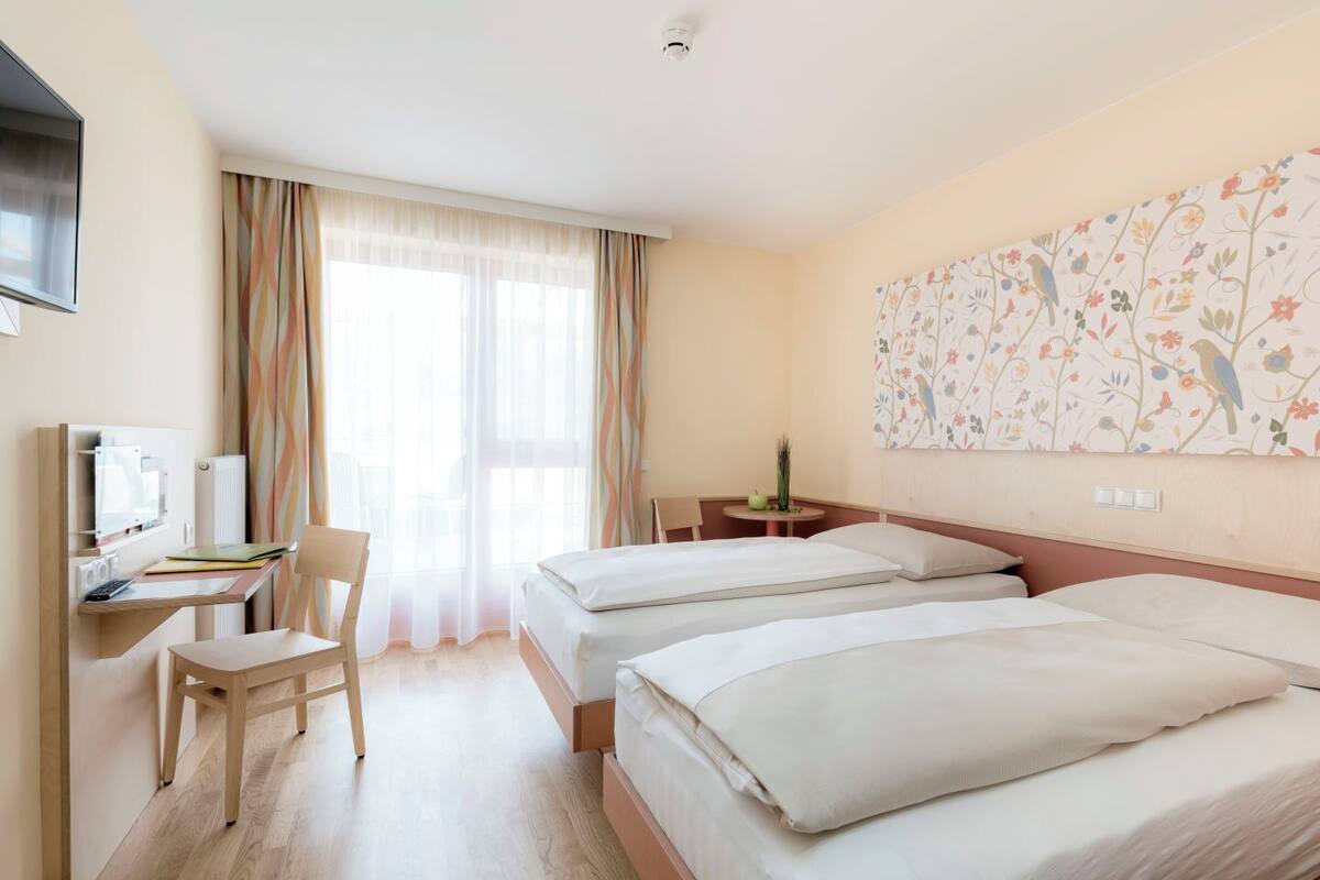 A hotel room with two beds, a desk with a chair, a TV, and a window with curtains.
