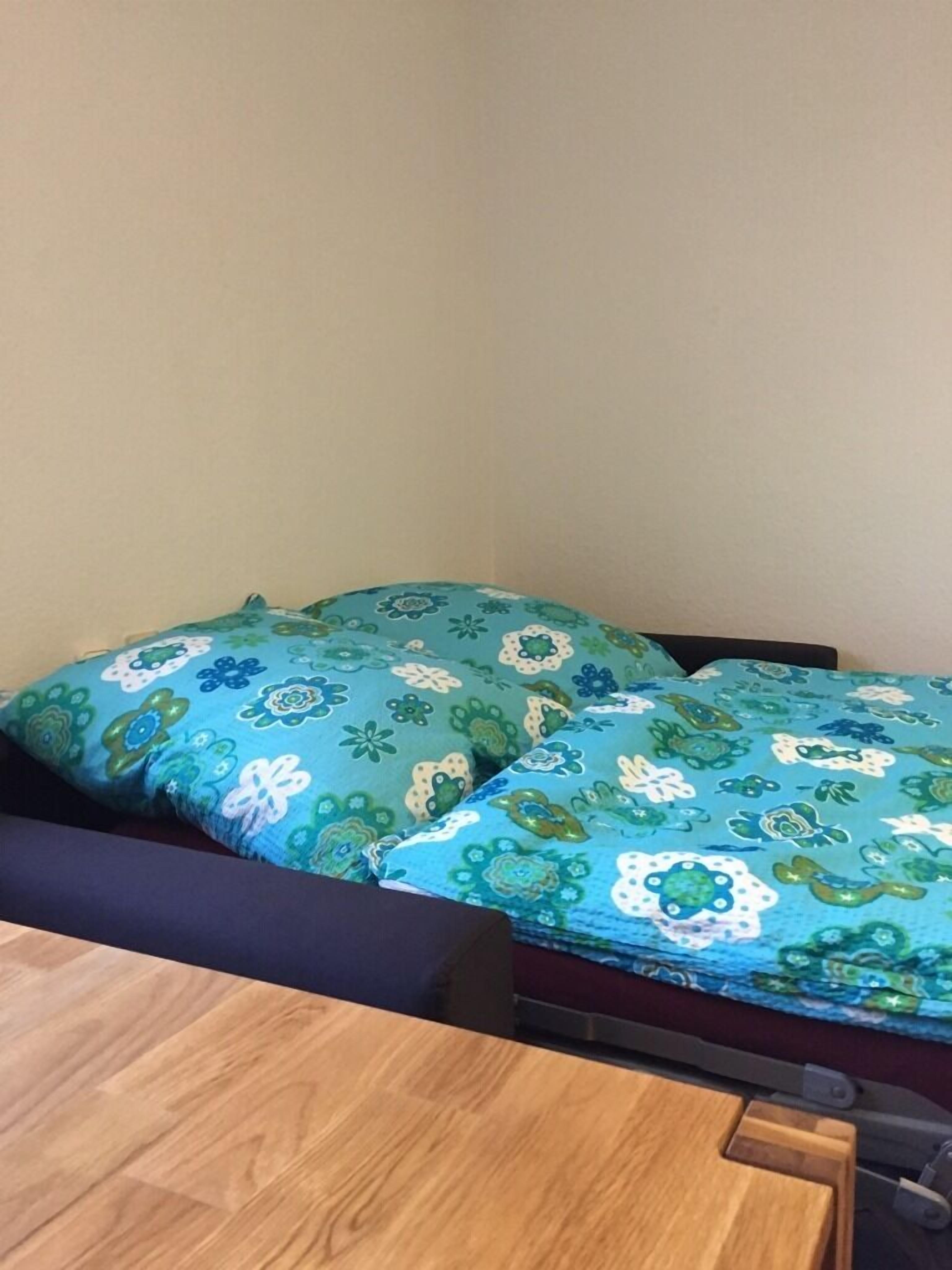 A room with a bed covered in floral-patterned bedding, a wooden table, and a chair.