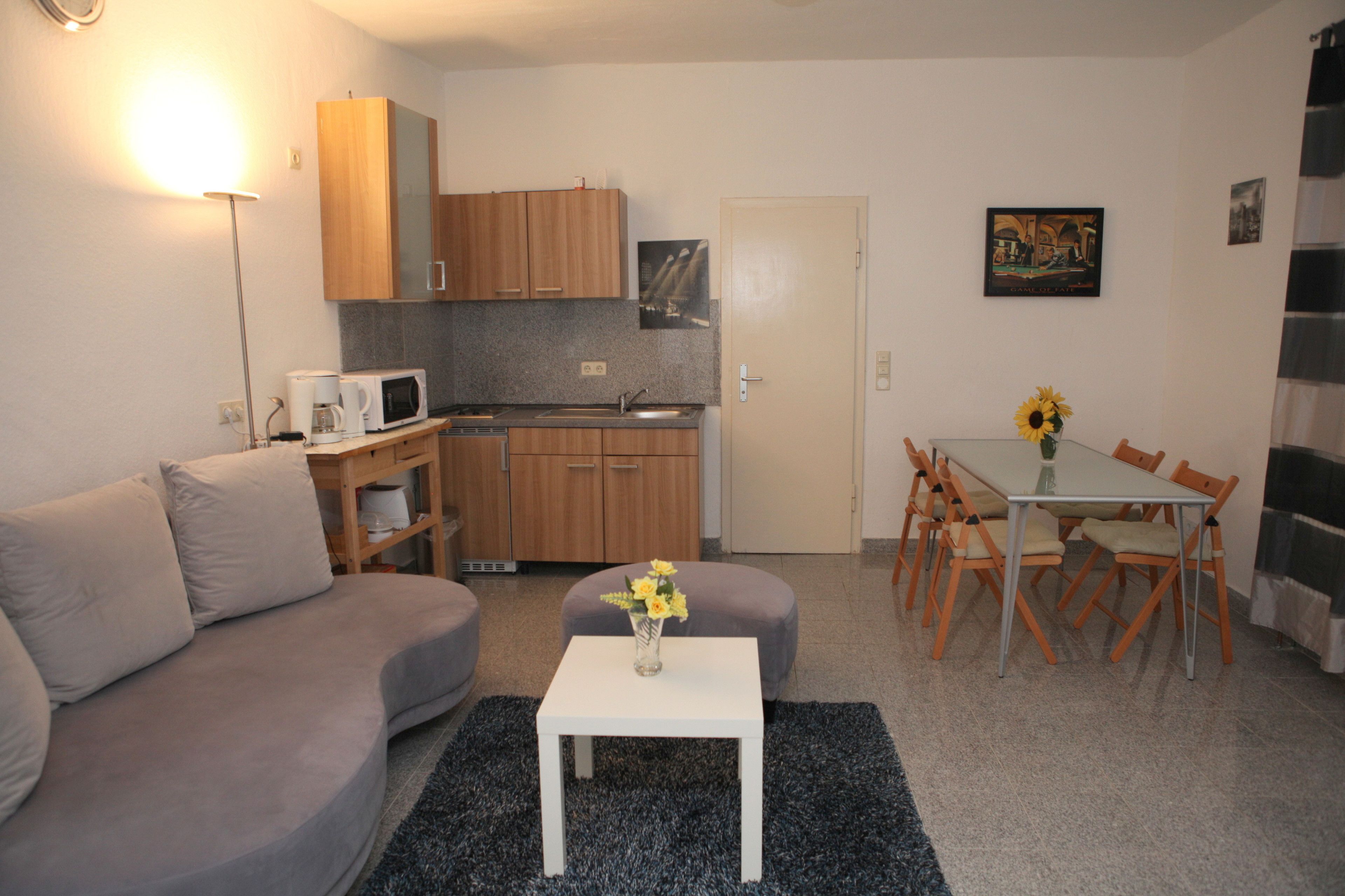 A compact living space with a kitchenette, a dining area with a table and chairs, a sofa, and a small white table with a vase.
