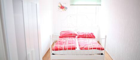 A small, white-framed bed with red bedding in a room with wooden flooring and white walls.