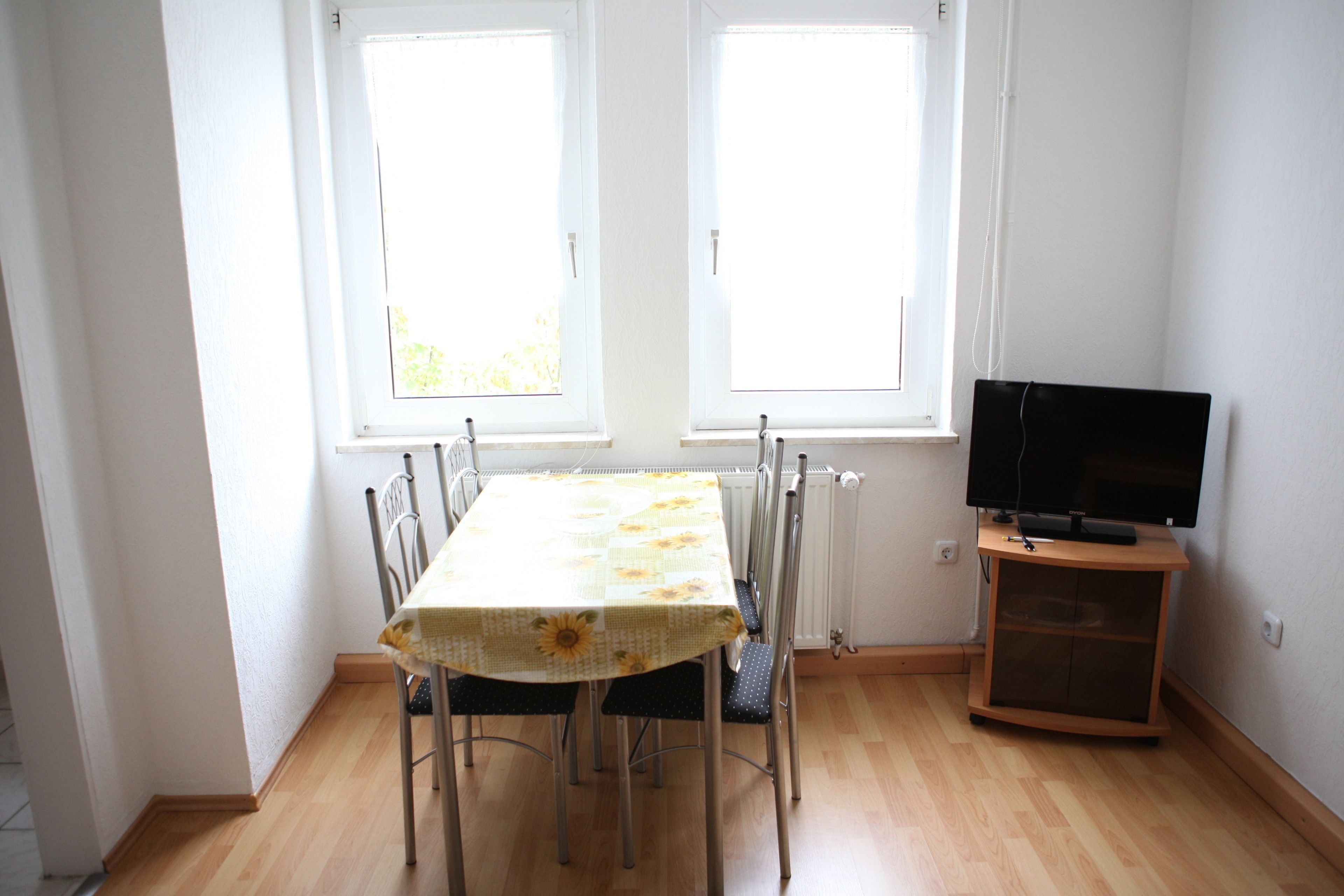 A dining area with a table and chairs, a television on a stand, and two windows with blinds.