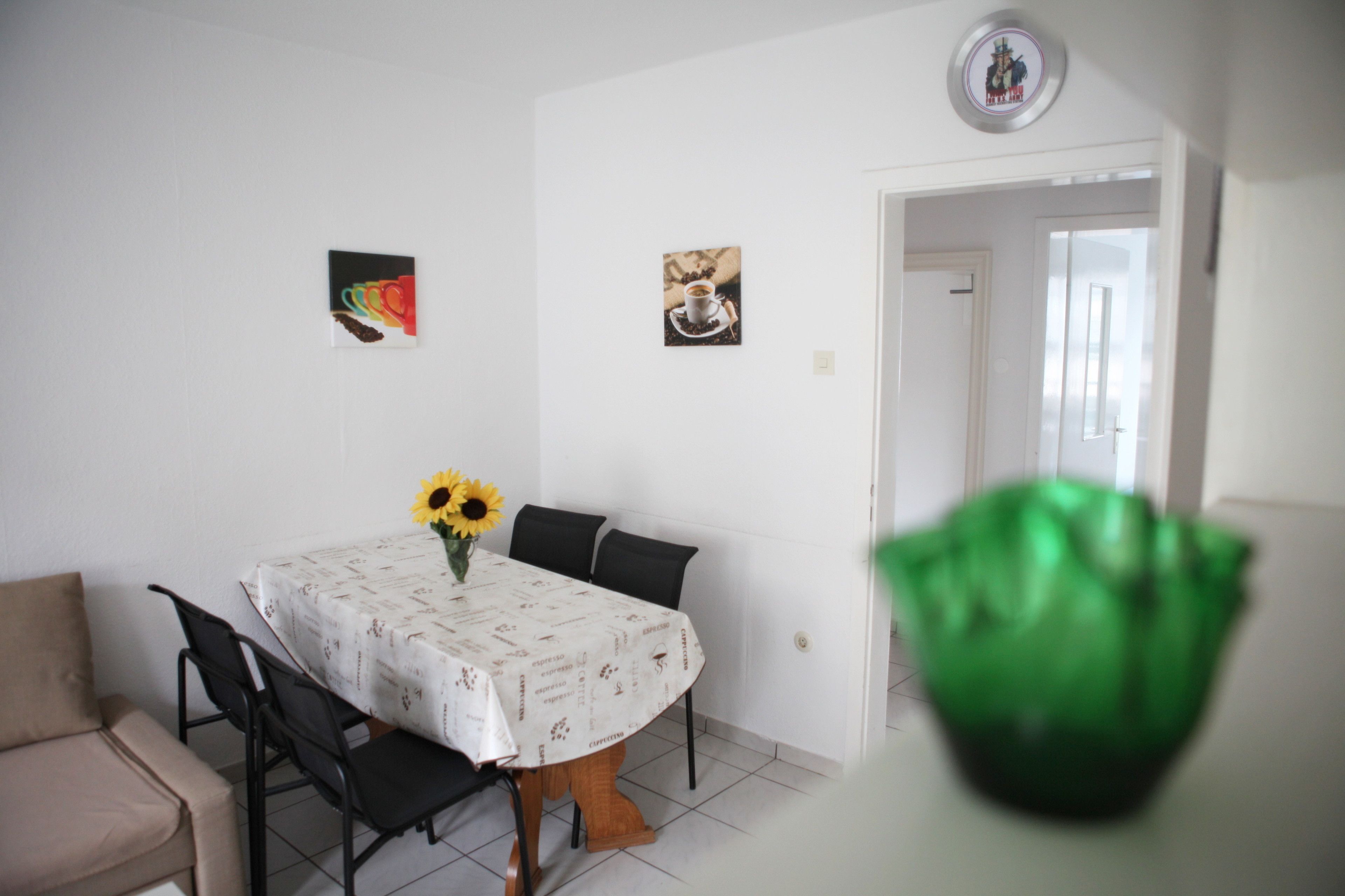 A dining area with a round table, chairs, and a vase with sunflowers. There are two framed pictures on the wall.