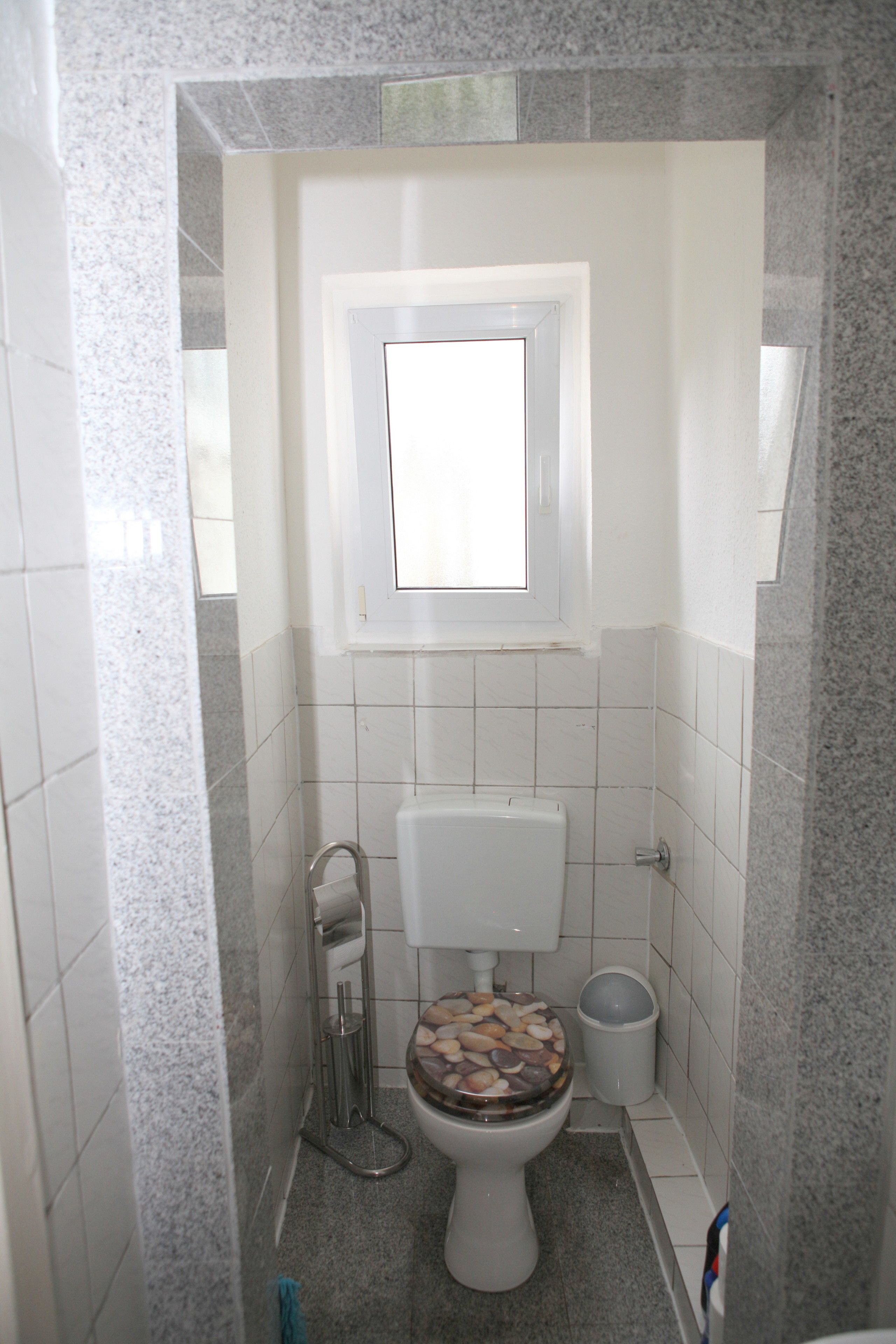 A small bathroom with a toilet, a window, and a trash bin.