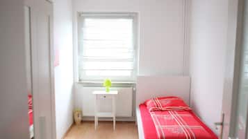 Une petite pièce lumineuse, avec un lit simple, un radiateur et une fenêtre équipée de stores.