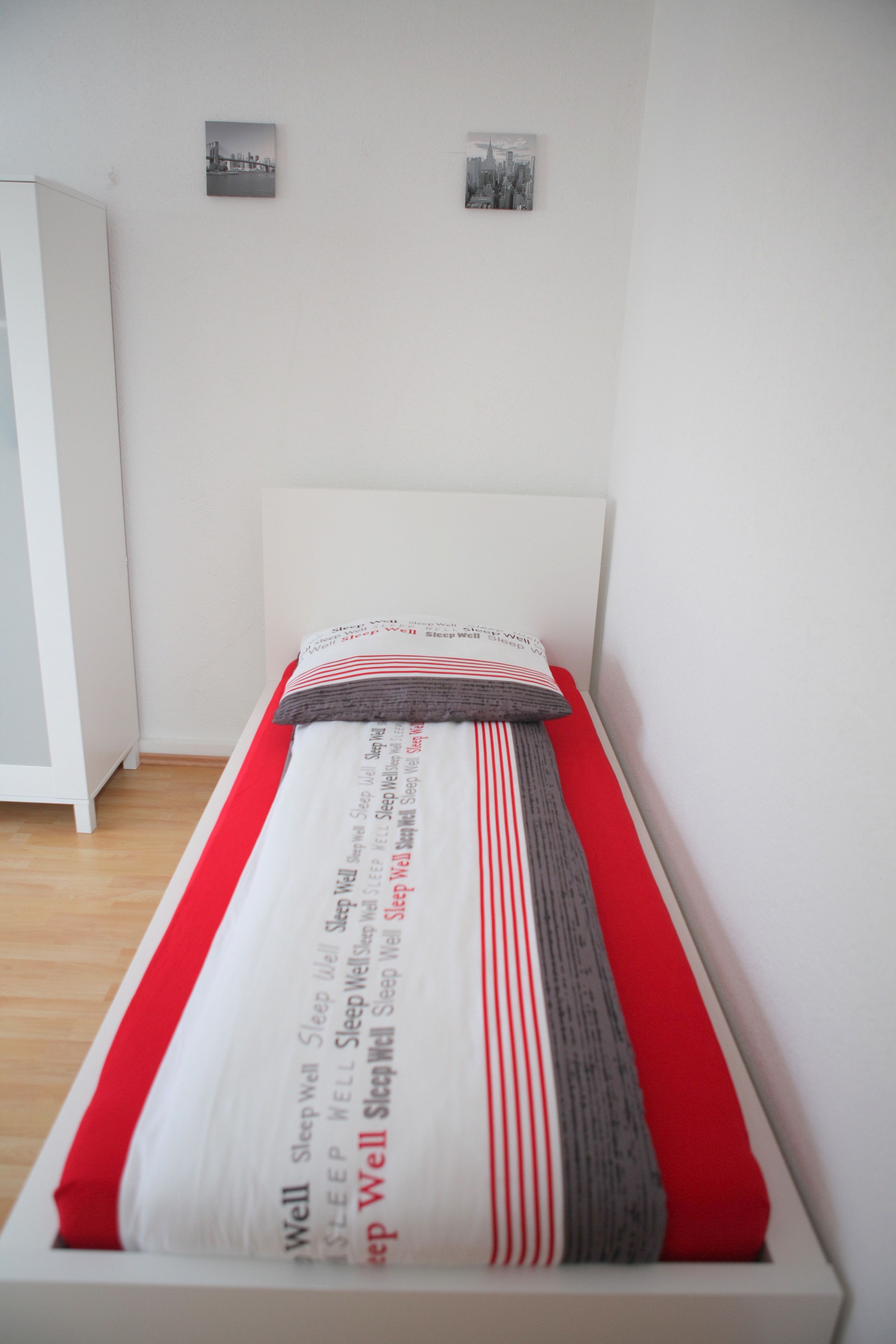 A neatly made bed with a white headboard, a red and white striped pillow, and a grey pillow. There are two small black and white pictures on the wall above the bed.