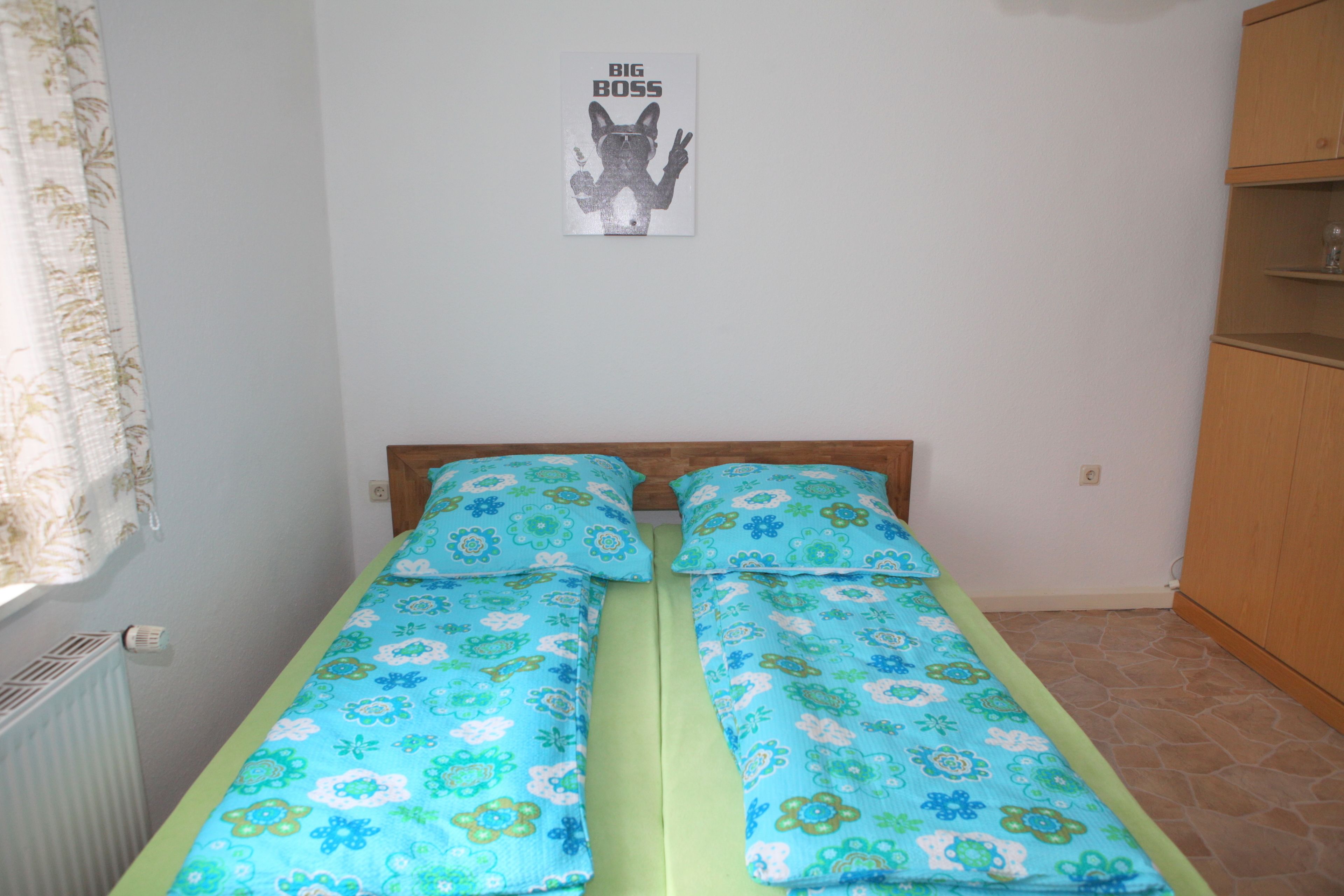Un lit simple avec une literie à motifs floraux, une tête de lit en bois, un radiateur, une affiche accrochée au mur et une armoire en bois.