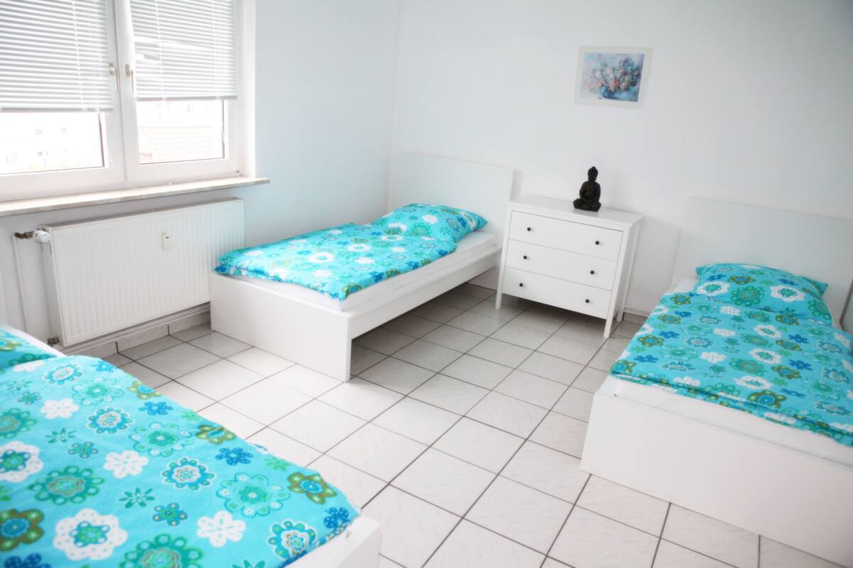 Een slaapkamer met twee eenpersoonsbedden, een witte dressoir, een radiator en een raam met zonwering.