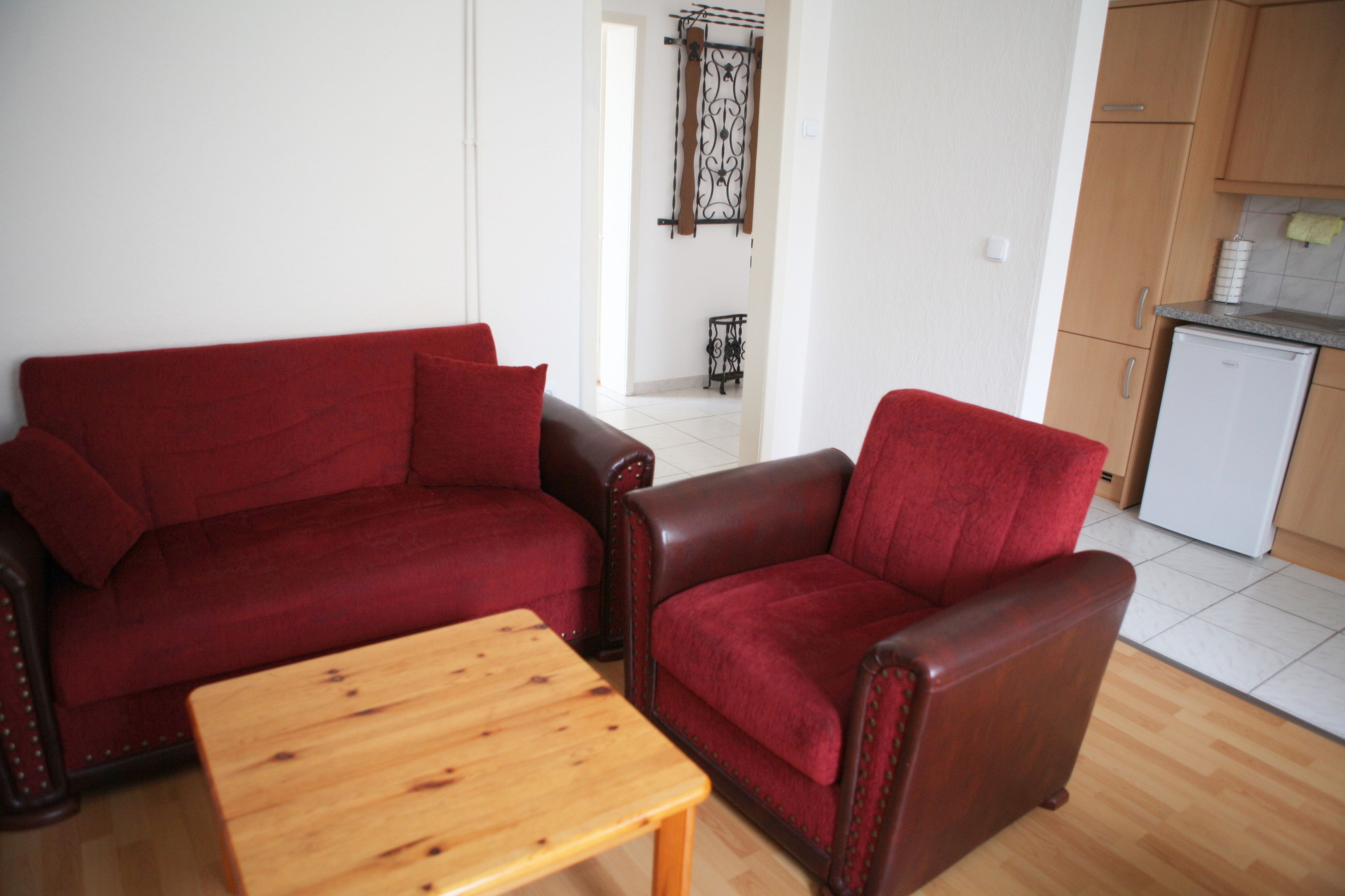 A living room with a red sofa, a wooden coffee table, and a kitchen area with wooden cabinets and a white refrigerator.