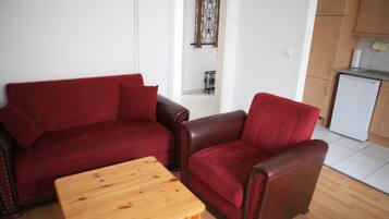 A living room with a red sofa, a wooden coffee table, and a kitchen area with wooden cabinets and a white refrigerator.