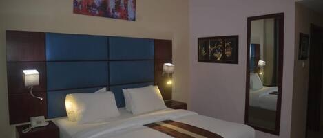 A hotel room with a large bed, a headboard with a blue panel, bedside lamps, a wall-mounted phone, and a mirror.