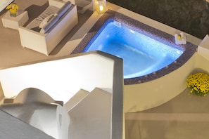 Un jacuzzi con borde de azulejos, rodeado por un área de descanso blanca con cojines y una pequeña mesa con flores.