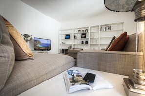 Un salon moderne comprenant un canapé, une table basse avec un magazine et un smartphone, un téléviseur et une étagère blanche ornée d’objets décoratifs.