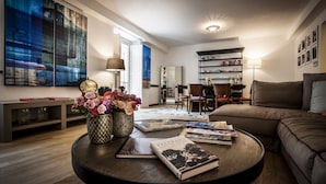 A modern living room with a round coffee table, books, and a vase of flowers. There is a sofa, a bookshelf, and a dining area in the background.