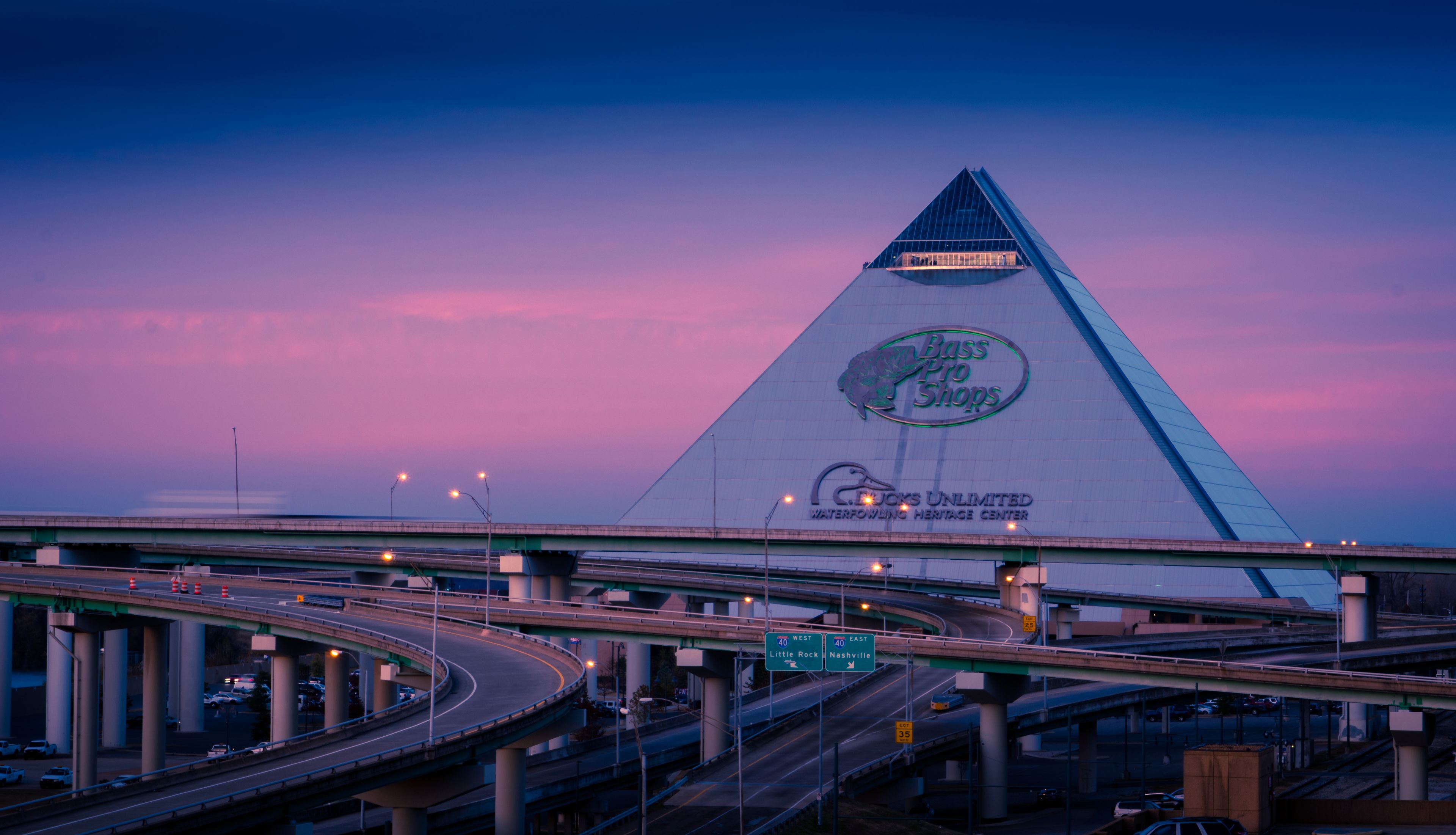 A highway interchange with multiple overpasses and a large building with a distinctive triangular roof and signage for 'Bass Pro Shops' and 'Waterpower Heritage Center'.