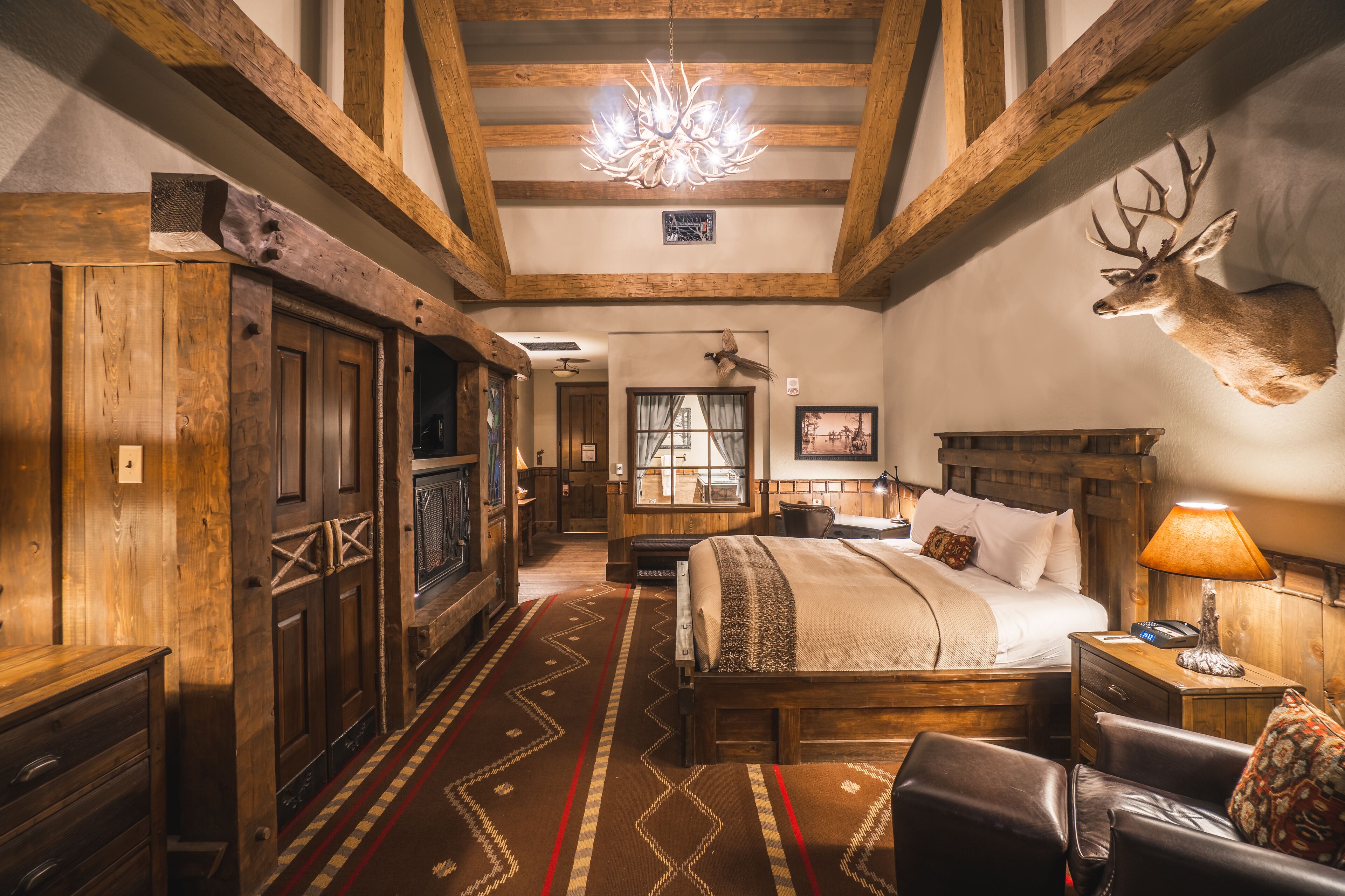 A rustic room with a wooden bed, a mounted deer head, a chandelier, and a traditional fireplace.