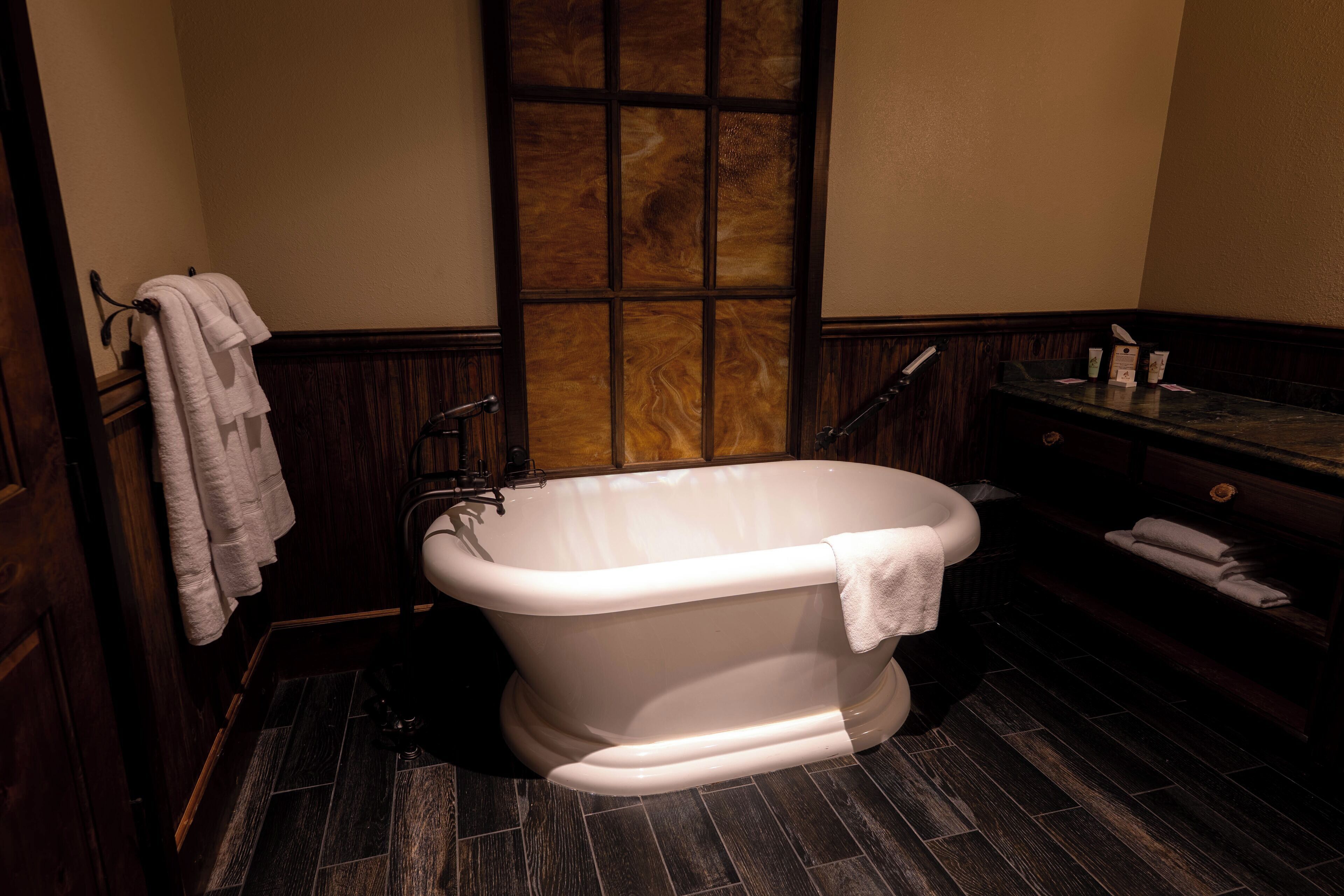 A classic clawfoot bathtub in a dimly lit bathroom with dark wood paneling and a large window.