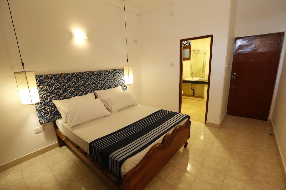 A neatly made bed with a patterned headboard, a bedside lamp, and a wooden door in a well-lit room with a tiled floor.