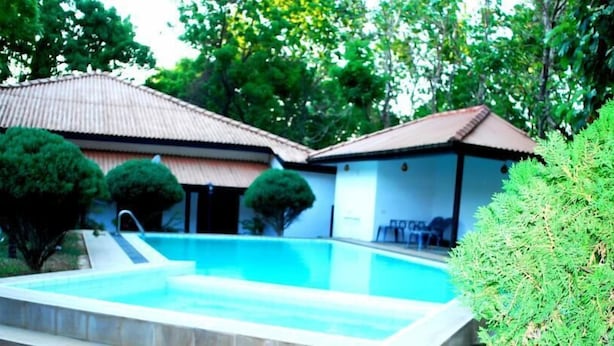 A swimming pool with a tiled deck, surrounded by greenery and a building with a tiled roof. - Jayasinghe Holiday Resort (Kataragama)