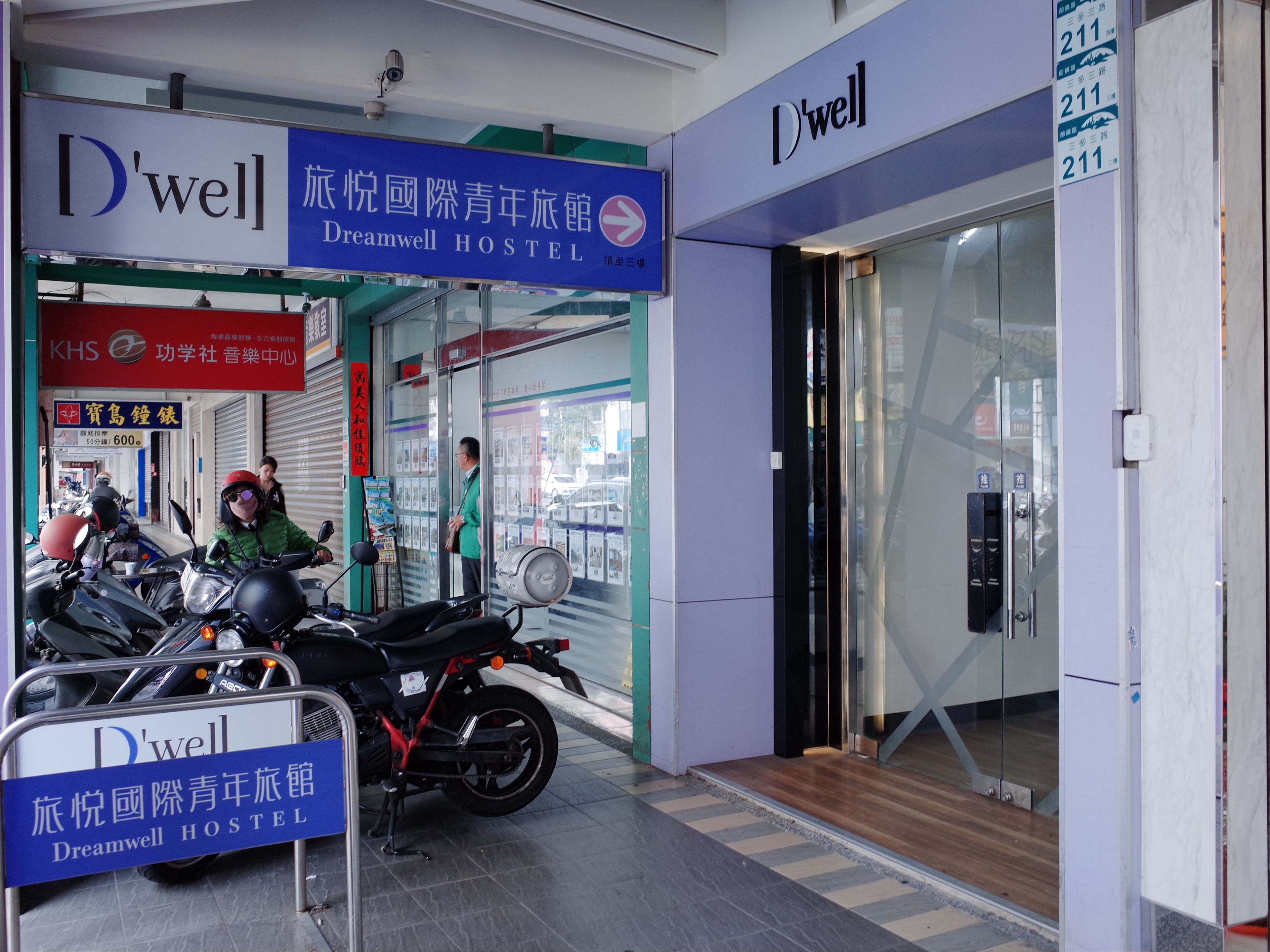 A Dreamwell Hostel entrance with a signboard, a man standing by the door, and several motorcycles parked outside.