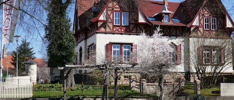 Una casa tradicional con entramado de madera, techo de tejas rojas, una chimenea de piedra y un pequeño jardín con flores y un árbol.