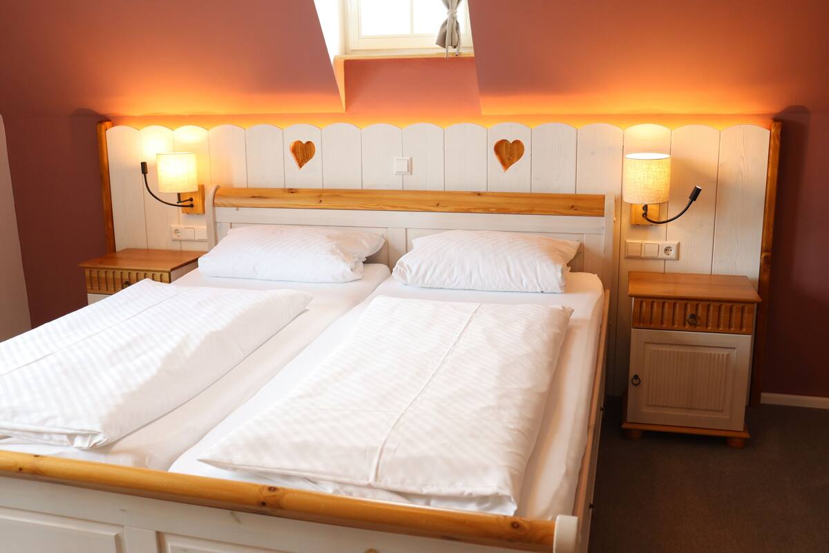 Een tweepersoonsbed met witte lakens, twee nachtkastjes en wandlampen met hartvormige uitsparingen.