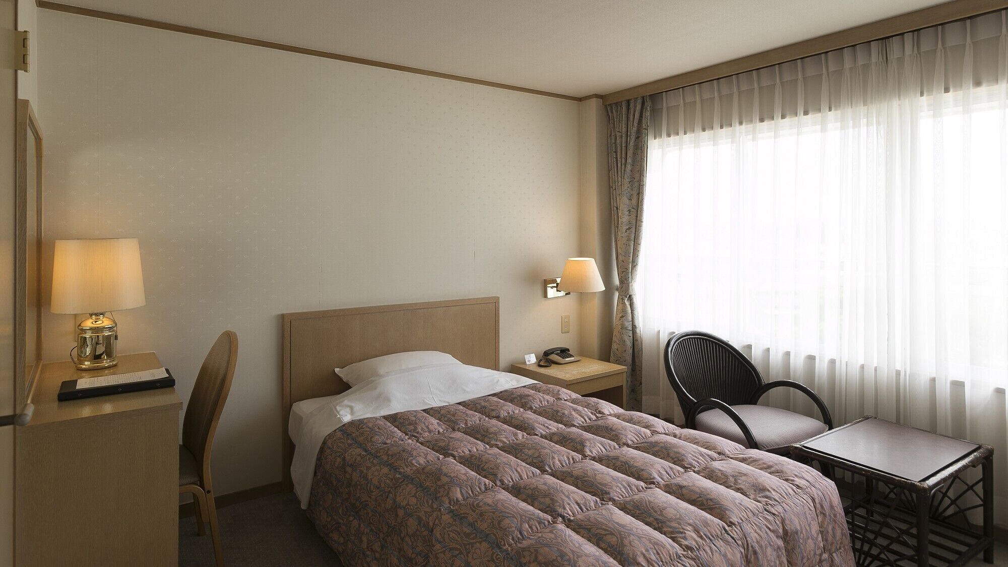 Een hotelkamer met een bed, bureau met lamp, stoel en tafel.