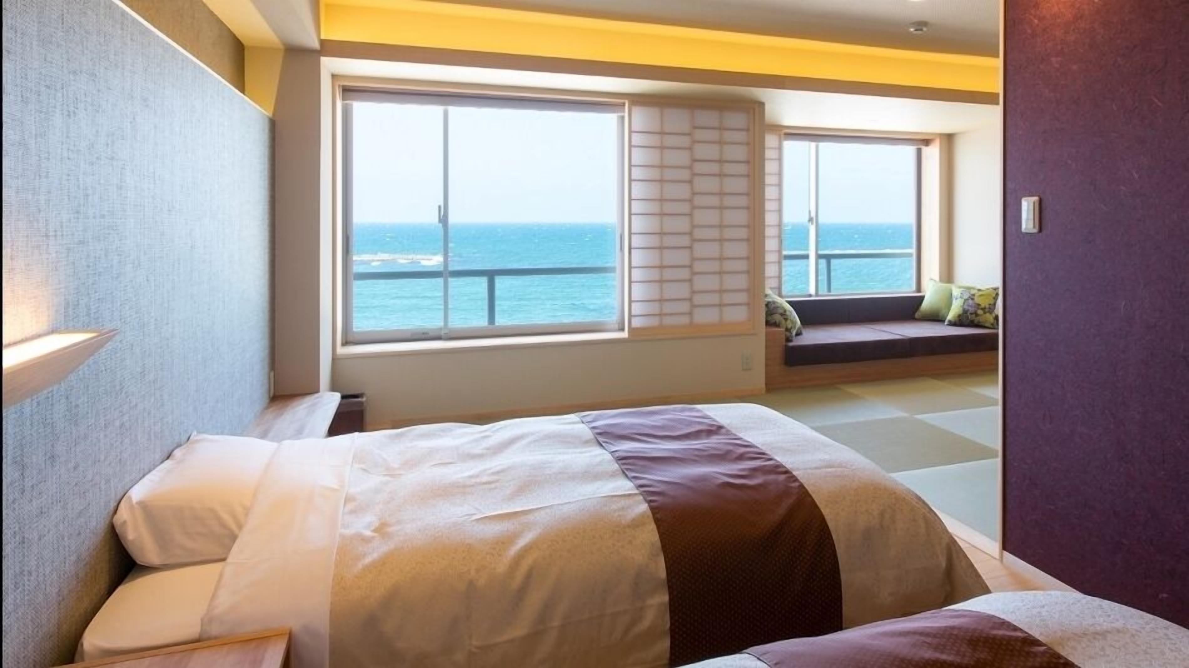 Een hotelkamer met een bed, een raam met uitzicht op zee, en een zithoek met kussens.