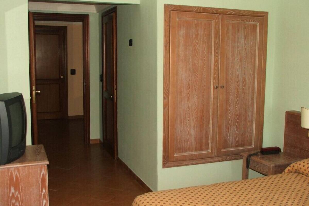A hotel room with a wooden wardrobe, a bed with patterned bedding, a bedside table with a lamp, and a television on a wooden stand.