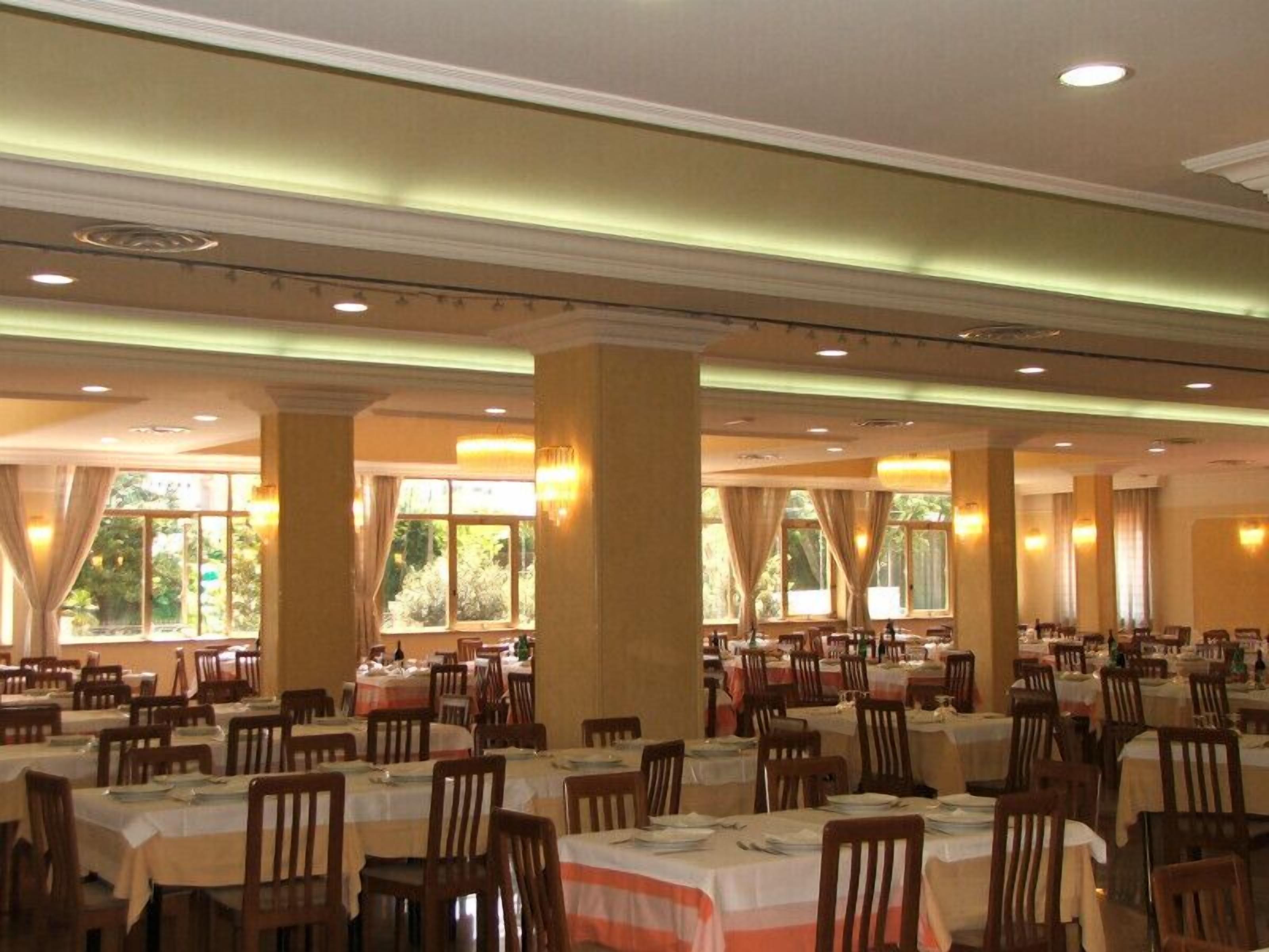 A large dining hall with multiple tables and chairs, high ceilings, and large windows allowing natural light.