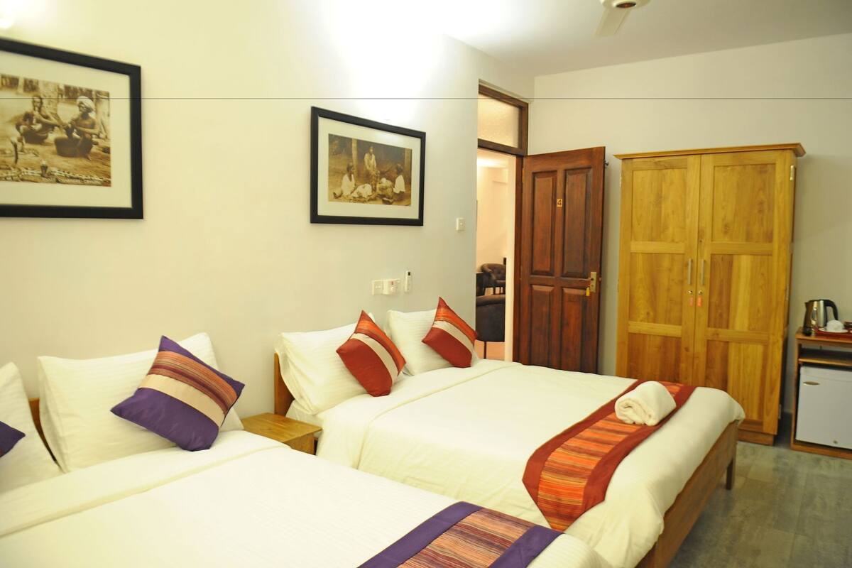 A hotel room with two beds, a wardrobe, a small kitchenette, and framed pictures on the wall.