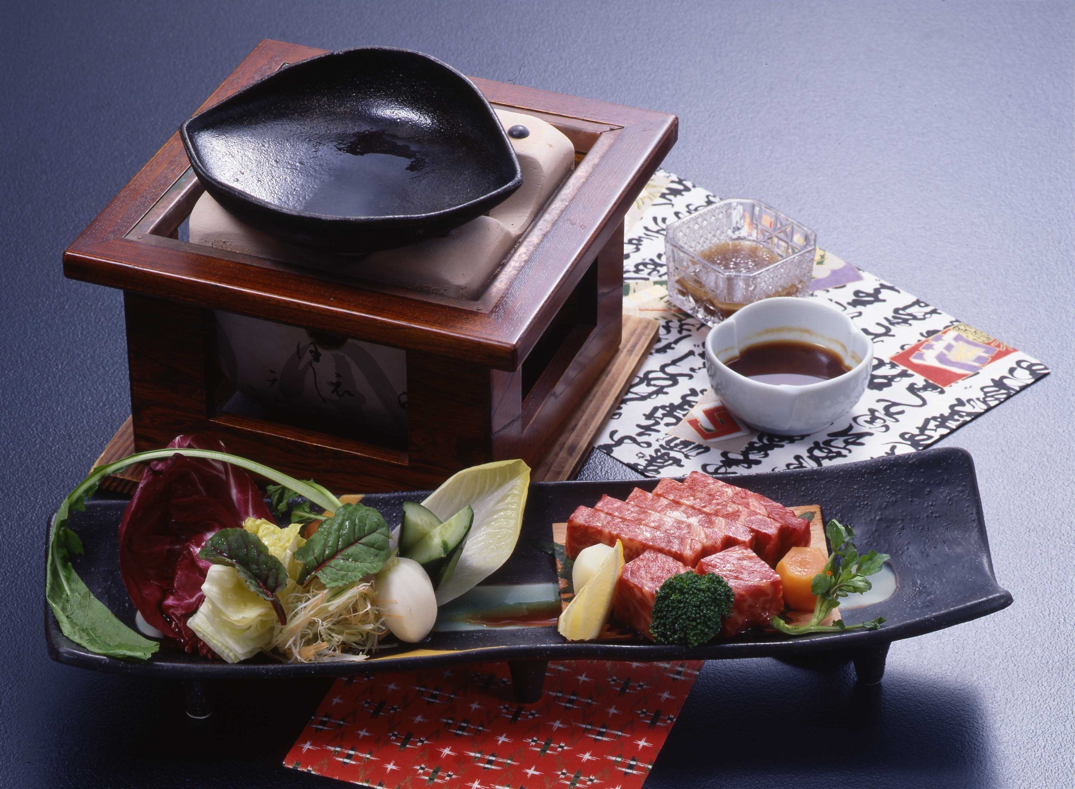 Una comida tradicional japonesa con una sartén negra, un tazón de salsa de soja y una variedad de verduras y carne en un plato negro.