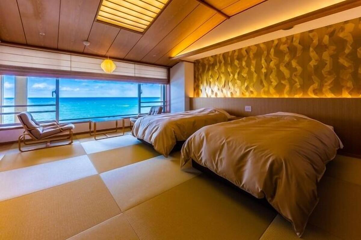 A room with two beds, a view of the sea, a lounge chair, and a patterned wall.